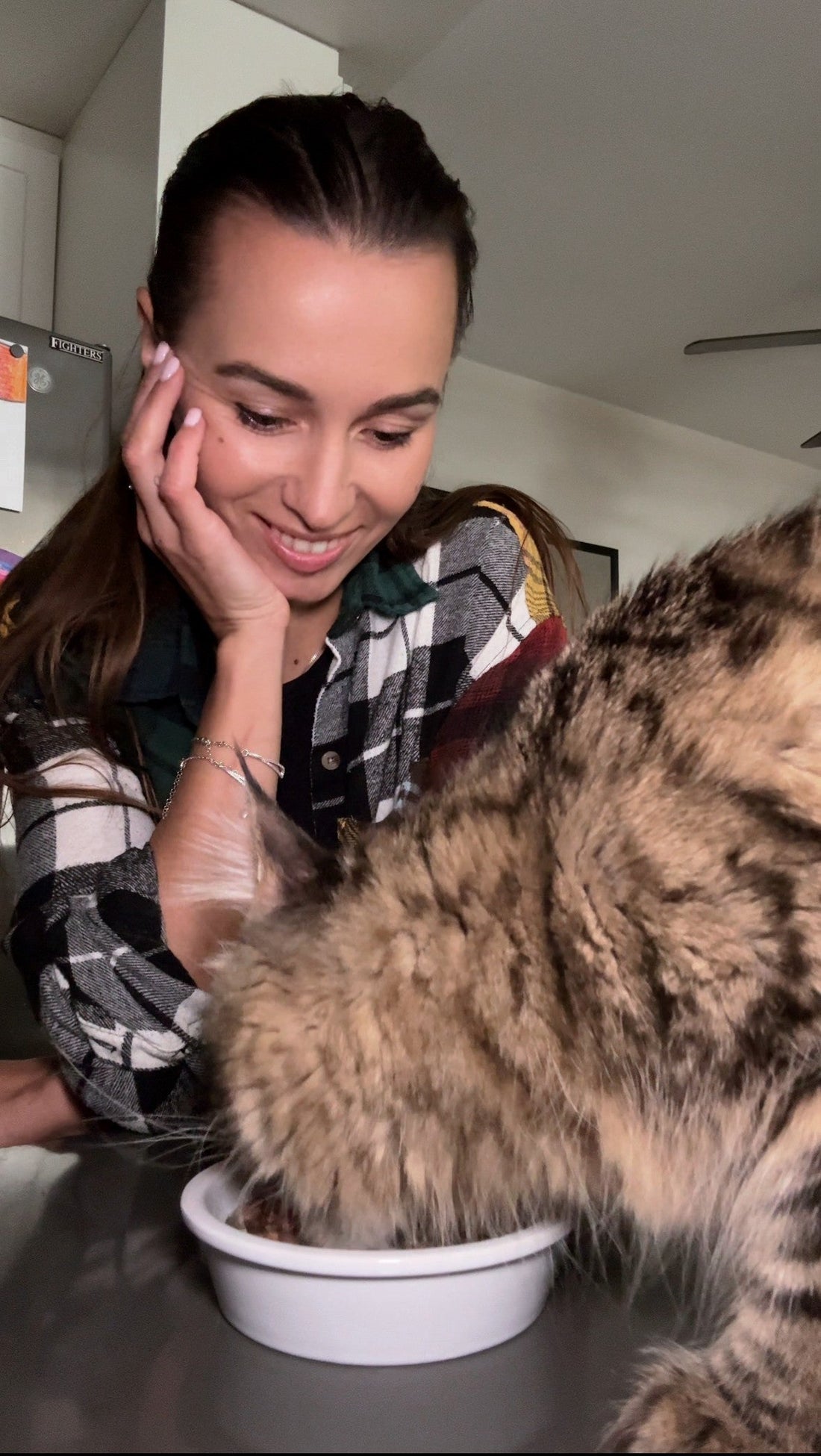 Alyona smiles while watching her Maine Coon cat Floppa eat from a ceramic food bowl on the table.
